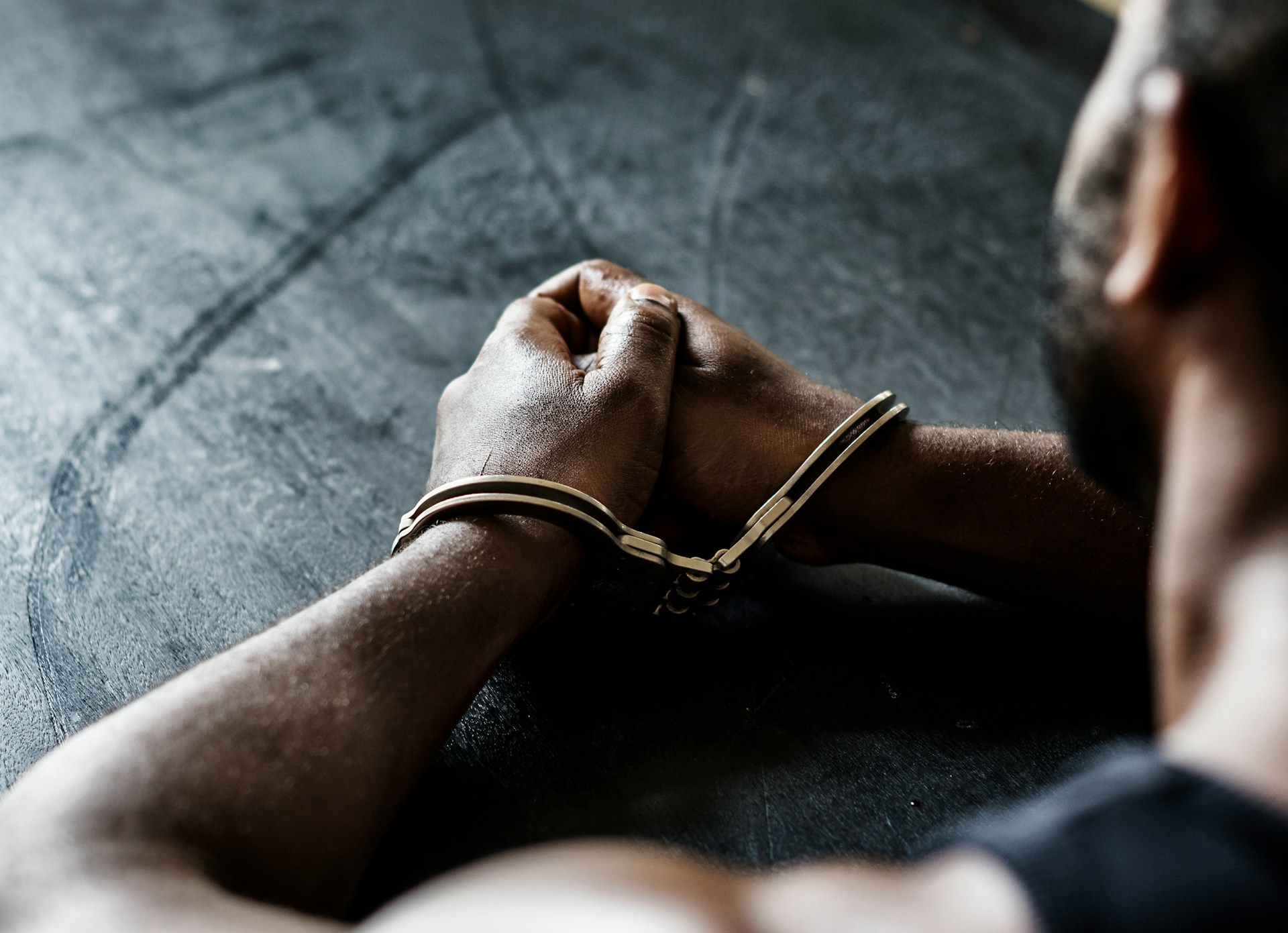 An arrested man with handcuffs clutching his hands together on a black table, the view set from behind his left shoulder and providing a small glimpse of the side of his face, representing the Good Samaritan Act immunity from arrest.