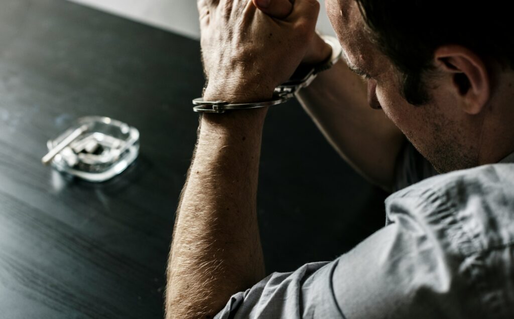A distressed man leans forward to rest his handcuffed hands with his elbows propped on a black table with an ashtray and smouldering cigarette, representing hate crimes.